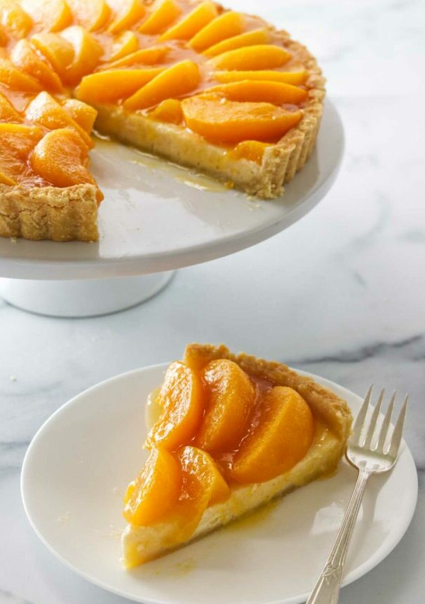 A peaches and cream tart on a cake pedestal in the background and a slice on a dessert plate with a fork in the foreground.