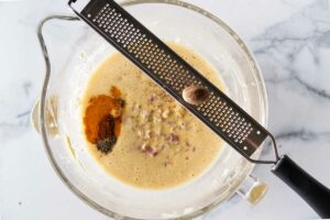 A mixing bowl with the ingredients for the ham and cheese strata. A microplane grating a nutmeg into the mixing bowl.