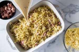 Pouring the cream custard over the ham and cheese strata. A dish of ham cubes in upper corner, a dish of shredded cheese in the lower corner.