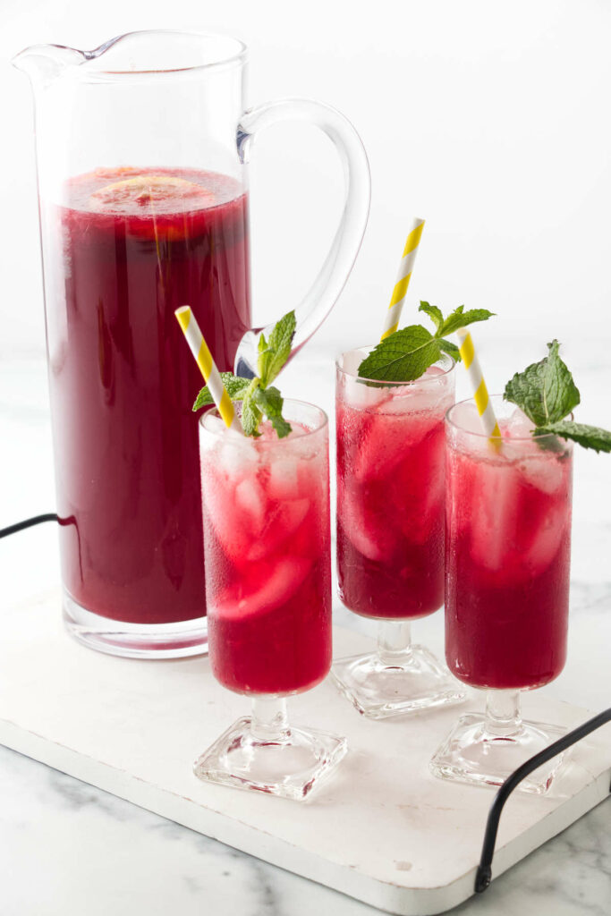 A serving tray with a pitcher filled with a red beverage and three glasses of red iced lemonade infused with hibiscus.