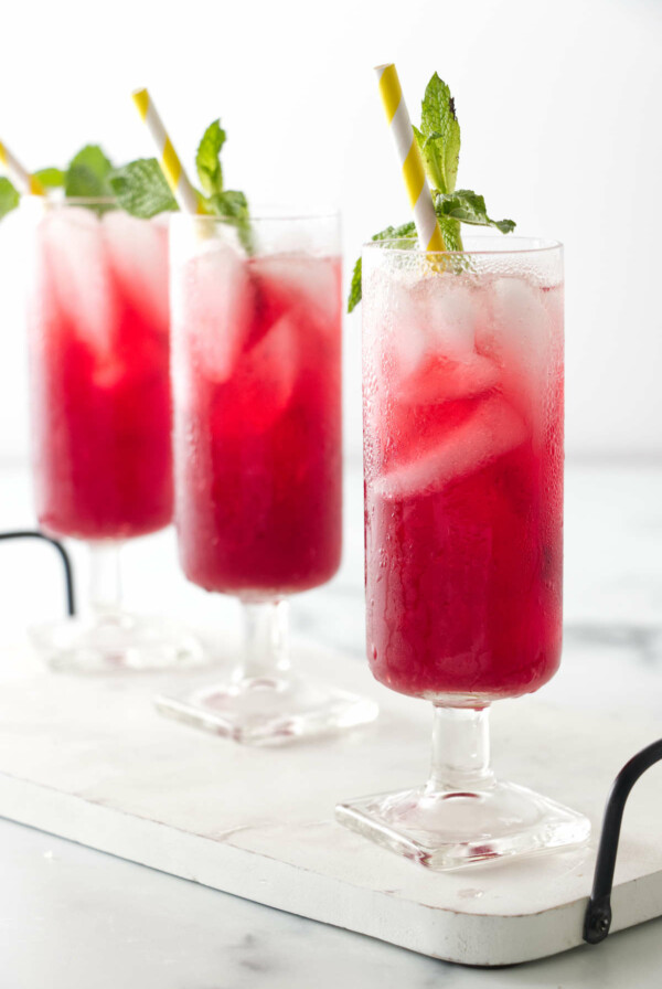 Three glasses of vibrant red lemonade made with hibiscus flowers, garnished with fresh mint leaves.