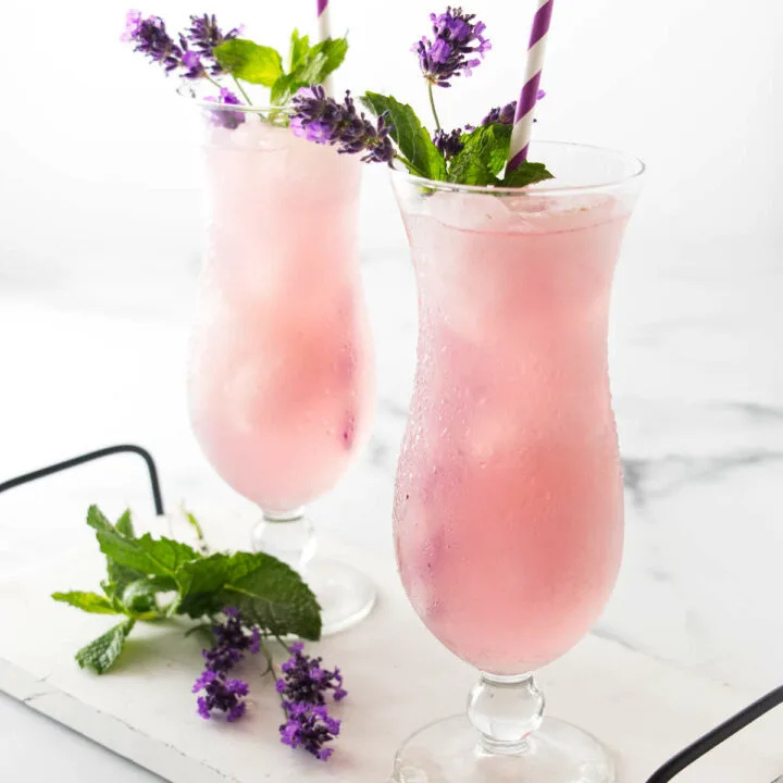 Two ice-filled glasses of lavender lemonade garnished with fresh lavender blooms, mint and with drinking straws.