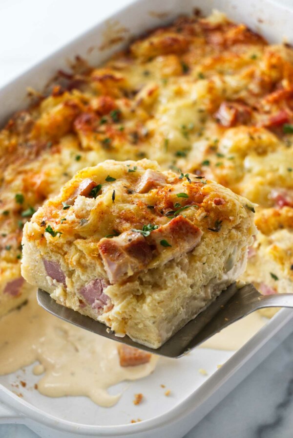 A spatula lifting a serving of ham and cheese strata from the casserole dish.