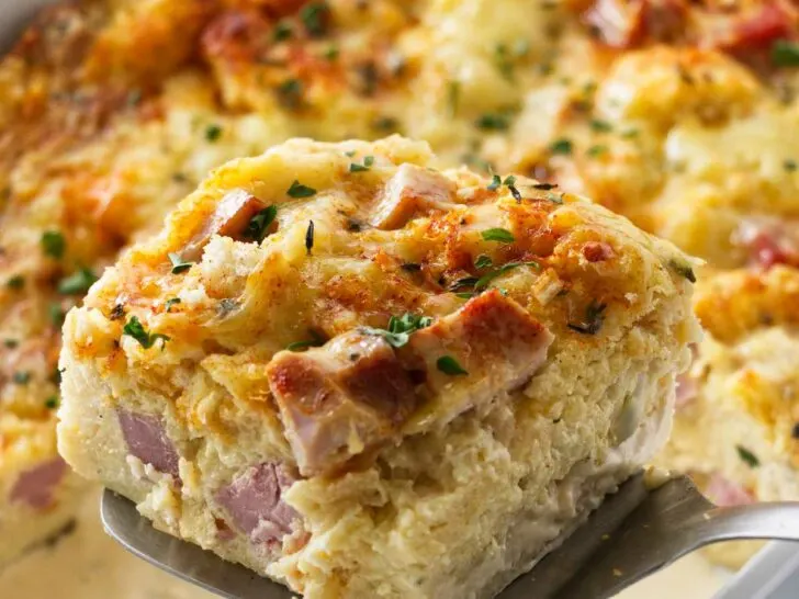 A spatula lifting a serving of ham and cheese strata from the casserole dish.
