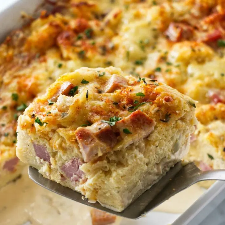 A spatula lifting a serving of ham and cheese strata from the casserole dish.