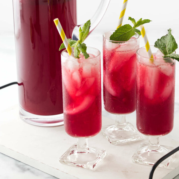 Three glasses of vibrant red hibiscus lemonade garnished with mint sprigs.next to a large pitcher filled with a red beverage.