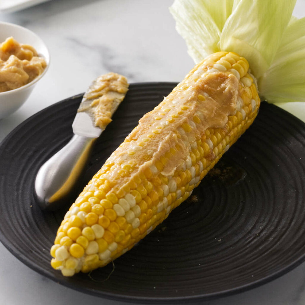 A serving of cooked corn on the cob on a plate with a smear of miso butter with a spreading knife. A small dish of miso butter to the left of the plate.