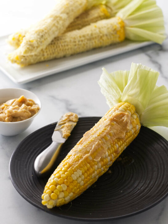 A serving of cooked corn on the cob smeared with miso butter and a spreading knife. A dish of miso butter to the left. A plate of corn cobs in the backgrround.