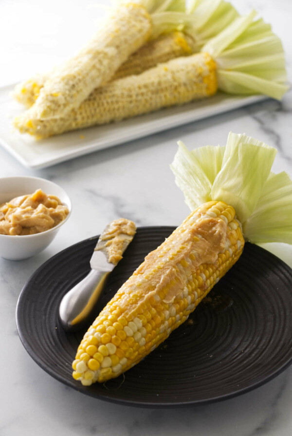 A serving of cooked corn on the cob smeared with miso butter and a spreading knife. A dish of miso butter to the left. A plate of corn cobs in the backgrround.