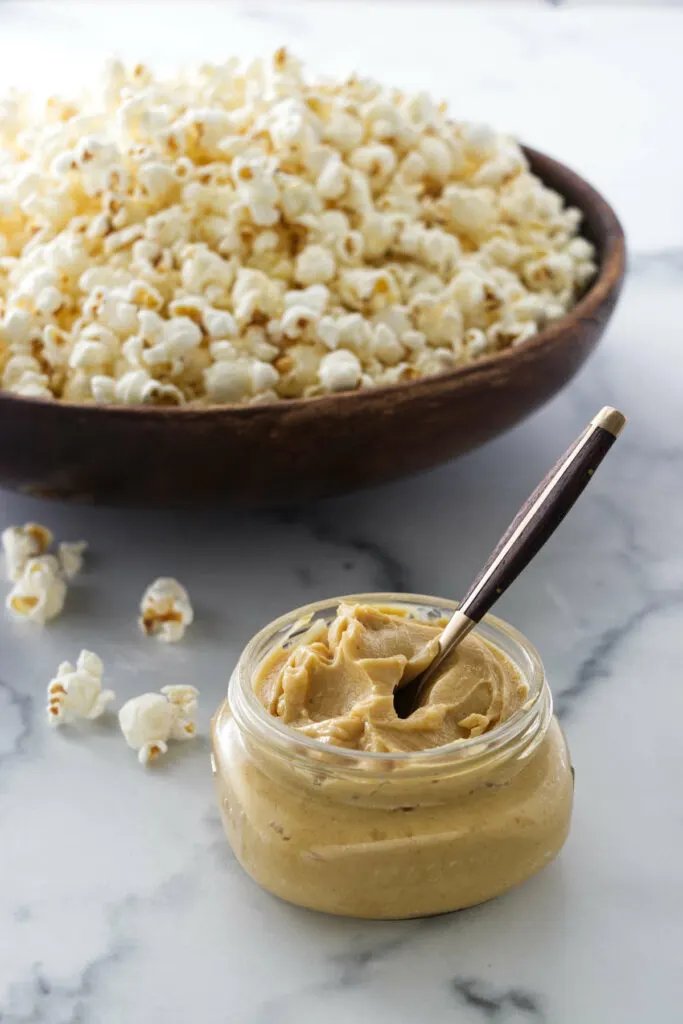 A jar of miso butter with a spoon and a wooden bowl of pop corn in the background.