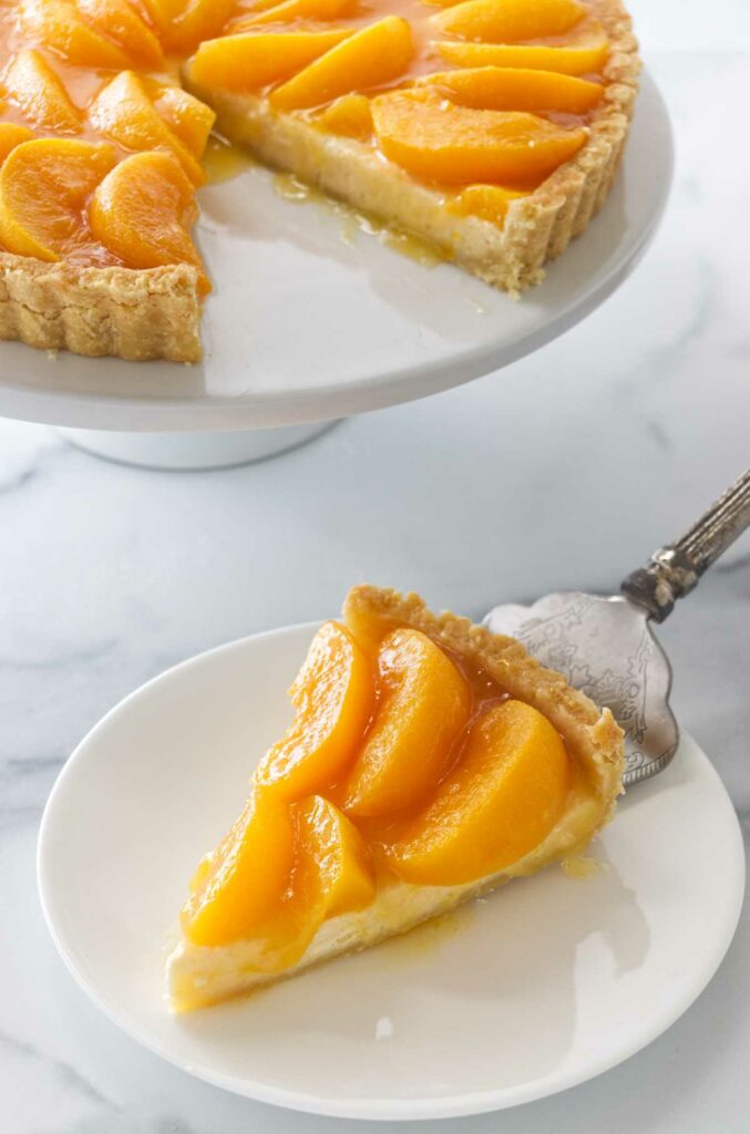 A spatula placing a slice of peaches and cream tart on a dessert plate. A cake pedestal with the tart in the background.