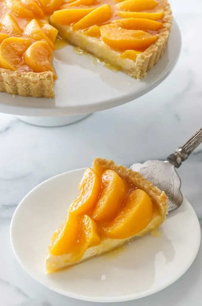 A spatula placing a slice of peaches and cream tart on a dessert plate. A cake pedestal with the tart in the background.