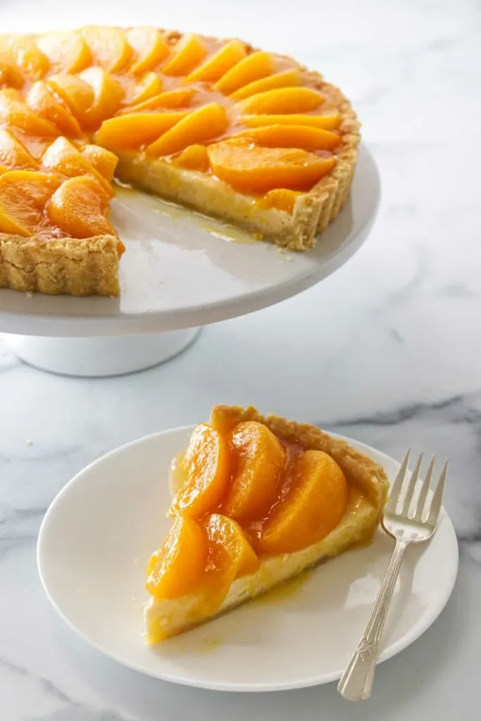 A peaches and cream tart on a cake pedestal in the background and a slice on a dessert plate with a fork in the foreground.