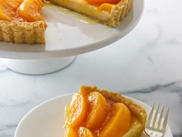 A peaches and cream tart on a cake pedestal in the background and a slice on a dessert plate with a fork in the foreground.