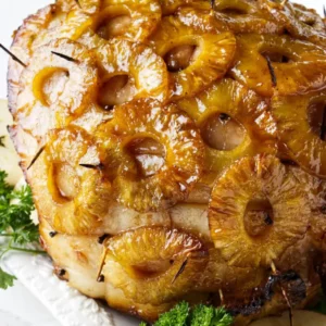 Glazed ham with pineapple rings, parsley, and apple slices on platter.
