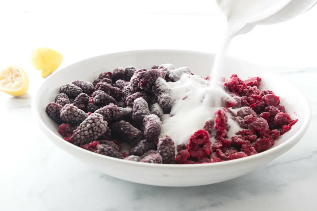 Pouring a sugar/thickener over a bowl of frozen fruit. Lemon rinds to the left of the bowl.