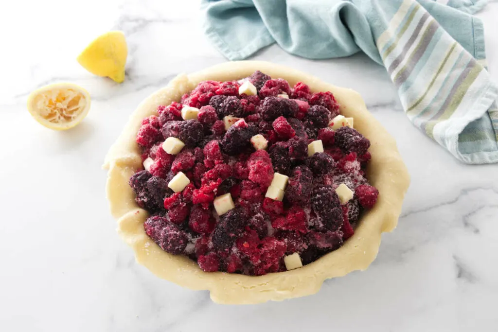 Raspberries, blackberries and butter bits in a pastry crust. Lemon rinds to the left of the pie and a kitchen towel in the background.