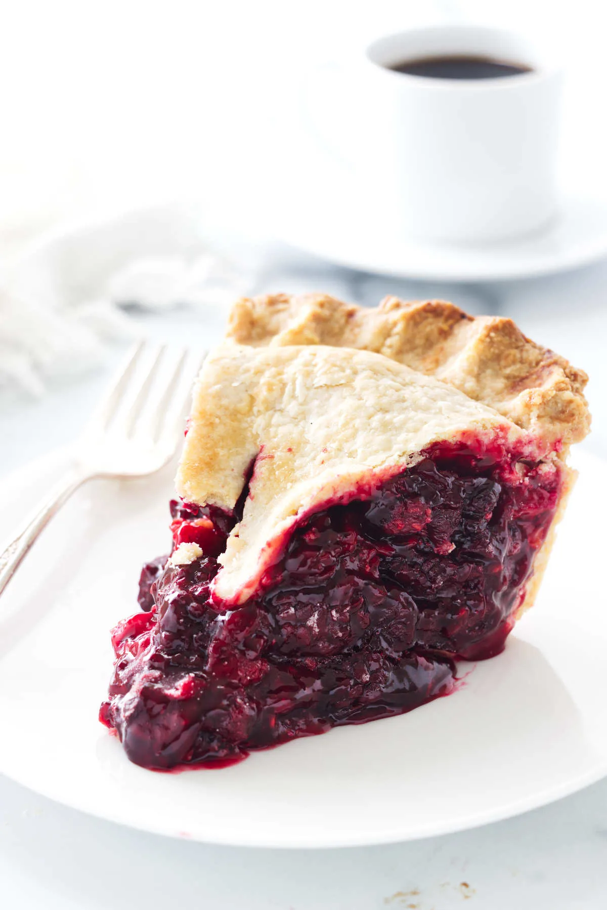 A serving of raspberry/blackberry pie on a plate and fork. Napkin and a cup of coffee in the background.