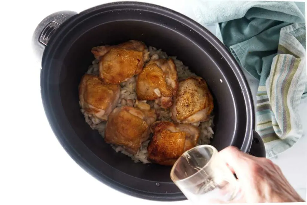 Pouring white wine into a slow cooker of browned chicken thighs.