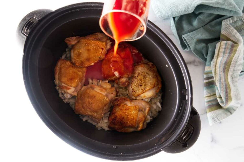 Pouring red tomato mixture over chicken thighs in a slow cooker.