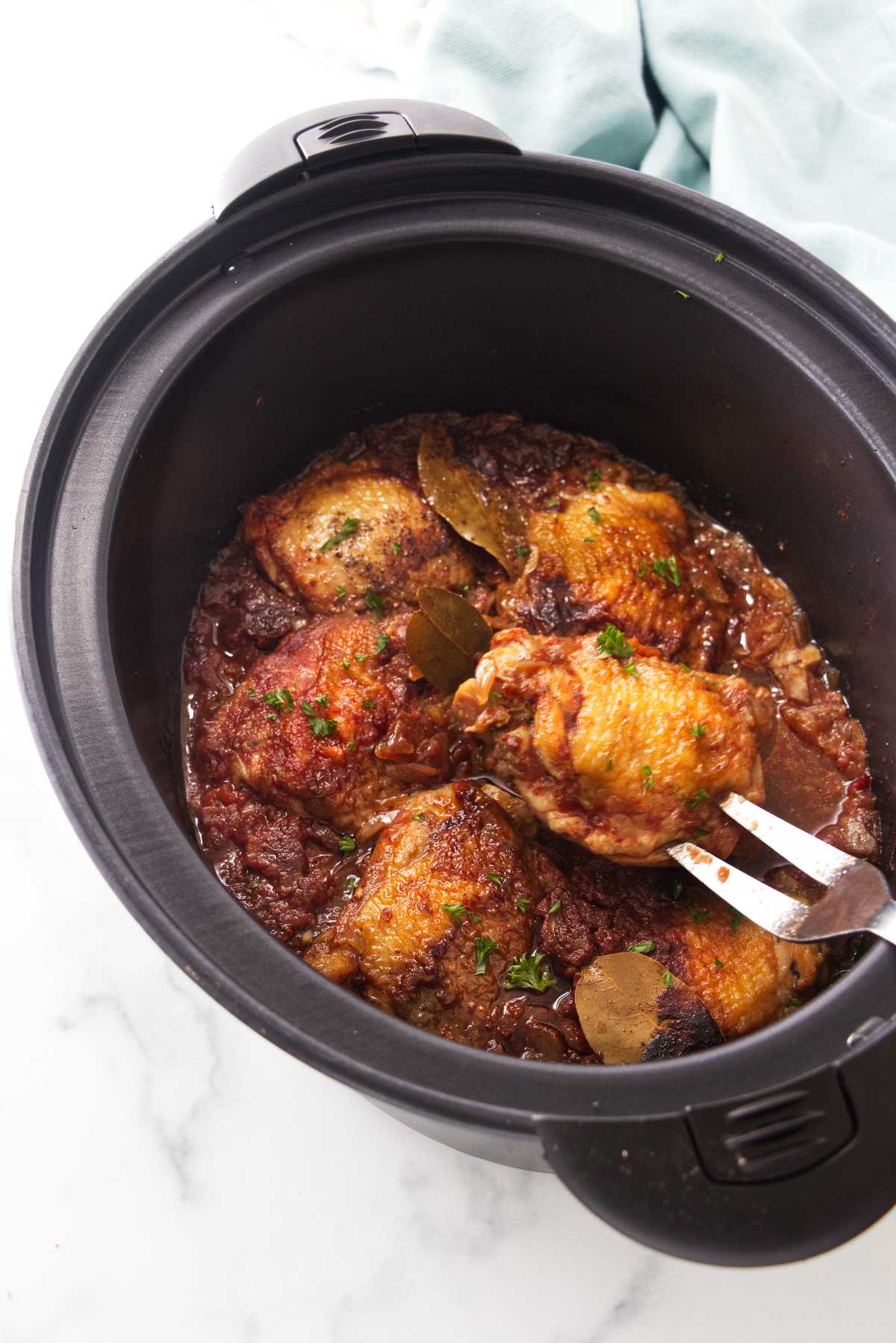 A slow cooker with chicken thighs in a paprika sauce. A large serving fork with one chicken thigh.
