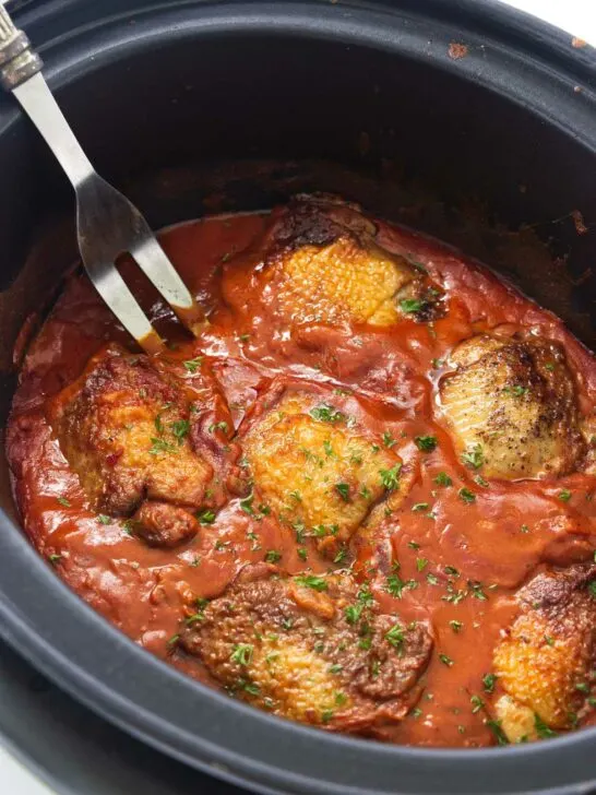 A slow cooker with chicken thighs in a paprika sauce and a large serving fork.