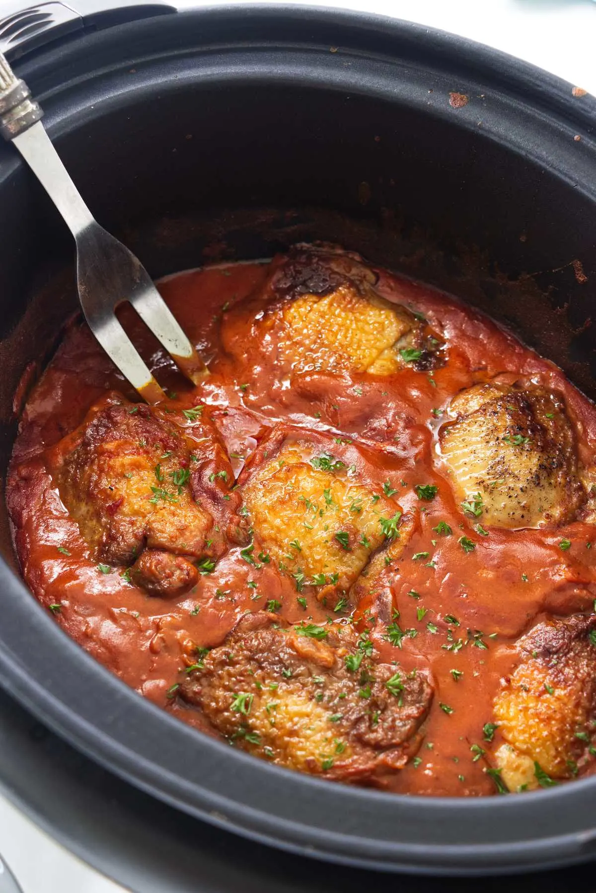 A slow cooker with chicken thighs in a paprika sauce and a large serving fork.