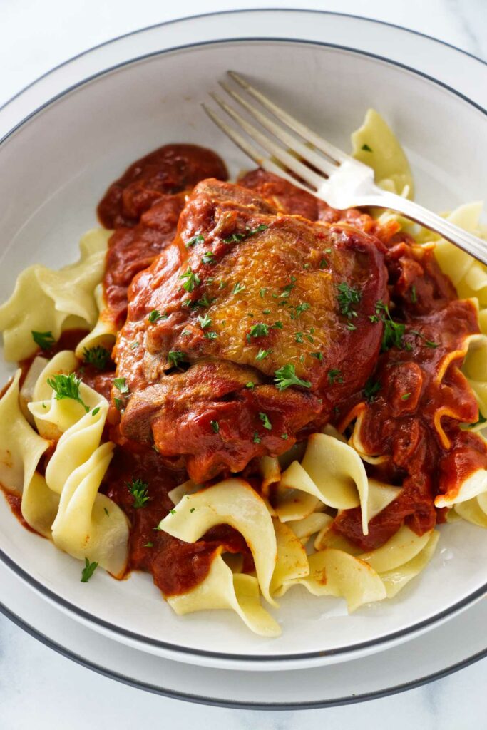 A dish with a cooked chicken thigh and red sauce on a bed of noodles.