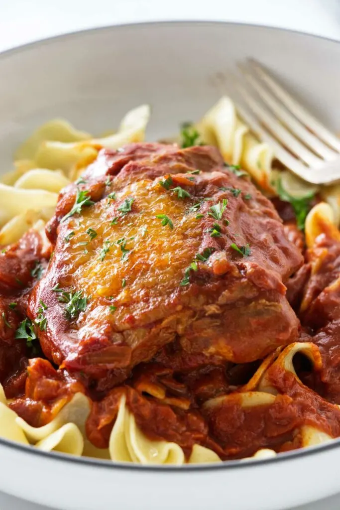 A dish with a cooked chicken thigh and red sauce on a bed of noodles.