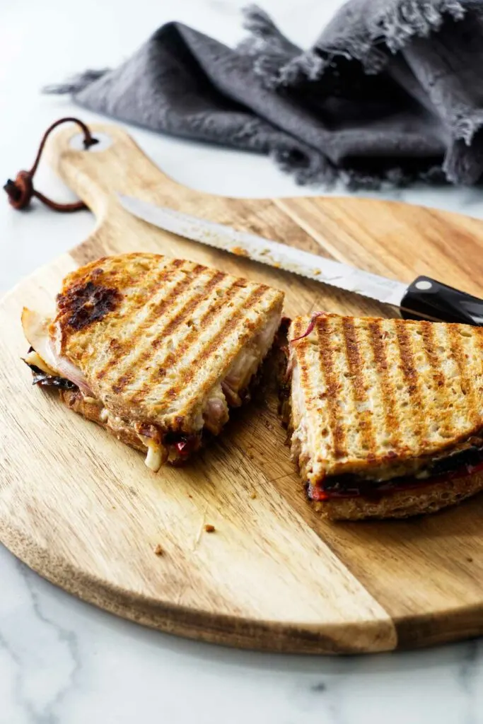 A panini sandwich cut in half on a wooden cutting board with a knife. A gray kitchen towel in the background.