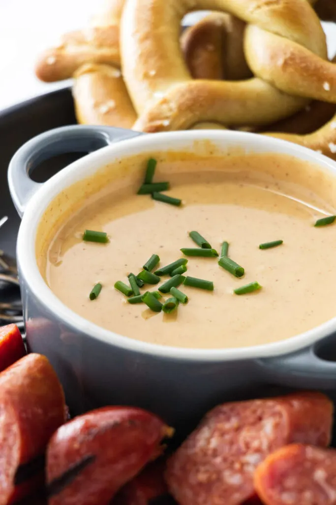 A small dish filled with melted cheese dip and garnished with chopped chives. Pretzels in the background, sausage chunks in the foreground.