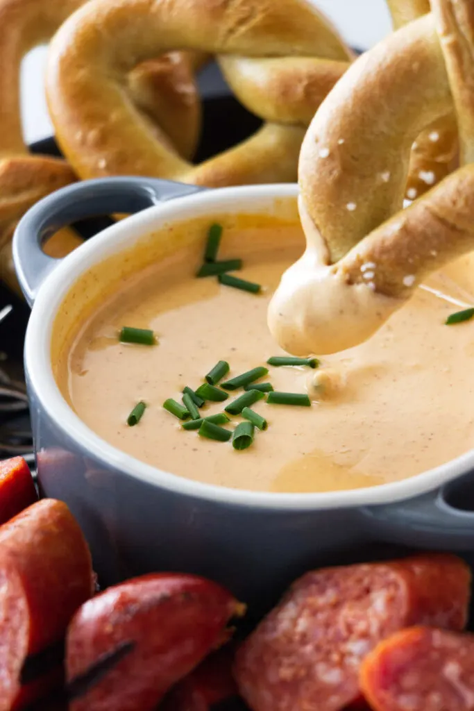 A dish of chive-garnished cheese dip with a pretzel being dipped, pretzels in the background and sausage chunks in the foreground.
