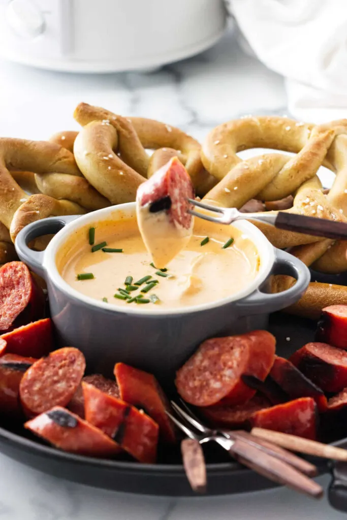 Party appetizer arrangement of a dish of dip and a small fork with sausage dipping into the dish. Pretzels and sausages included in the plate.