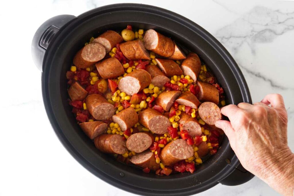 Adding sausages to the ingredients in a slow cooker.