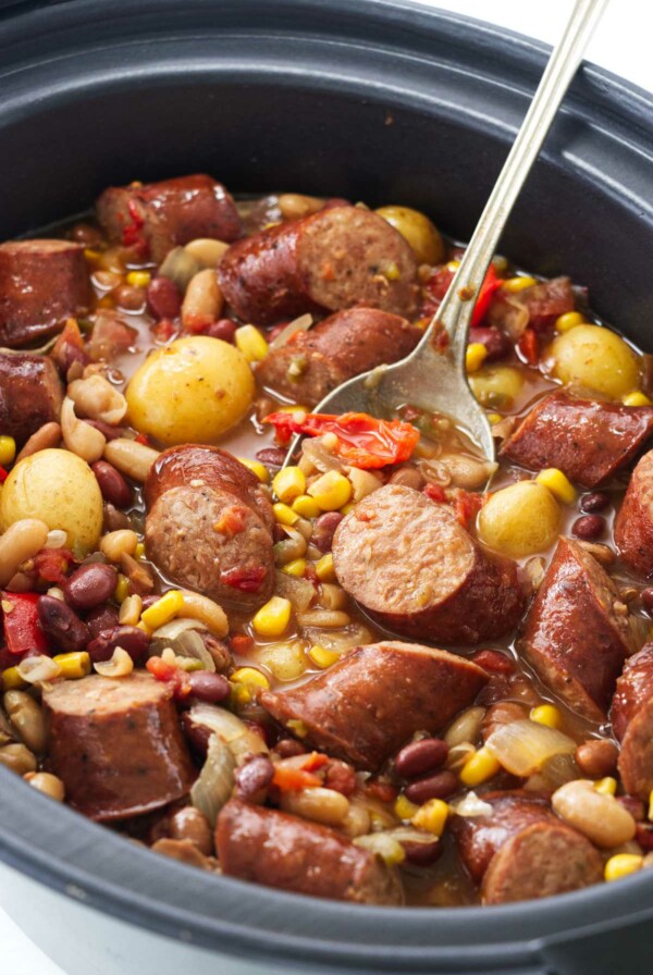 A crock pot with vegetables, sausage and a serving spoon.