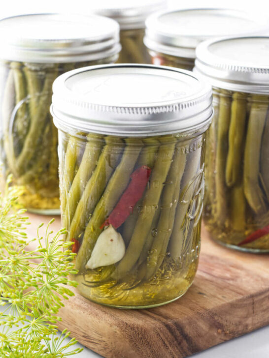 Pickled green beans in a canning jar with garlic and hot pepper, sitting on a cutting board with fresh dill in the corner.