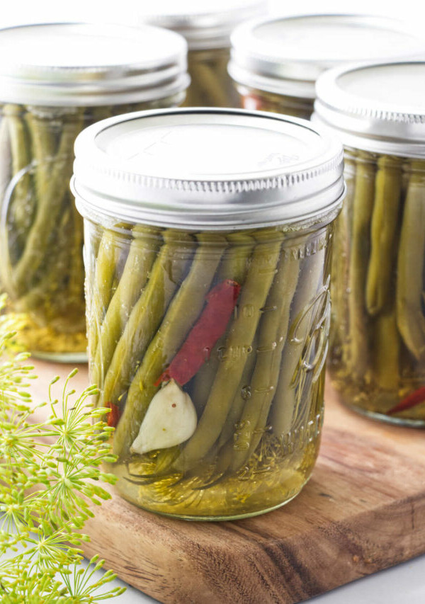 Pickled green beans in a canning jar with garlic and hot pepper, sitting on a cutting board with fresh dill in the corner.