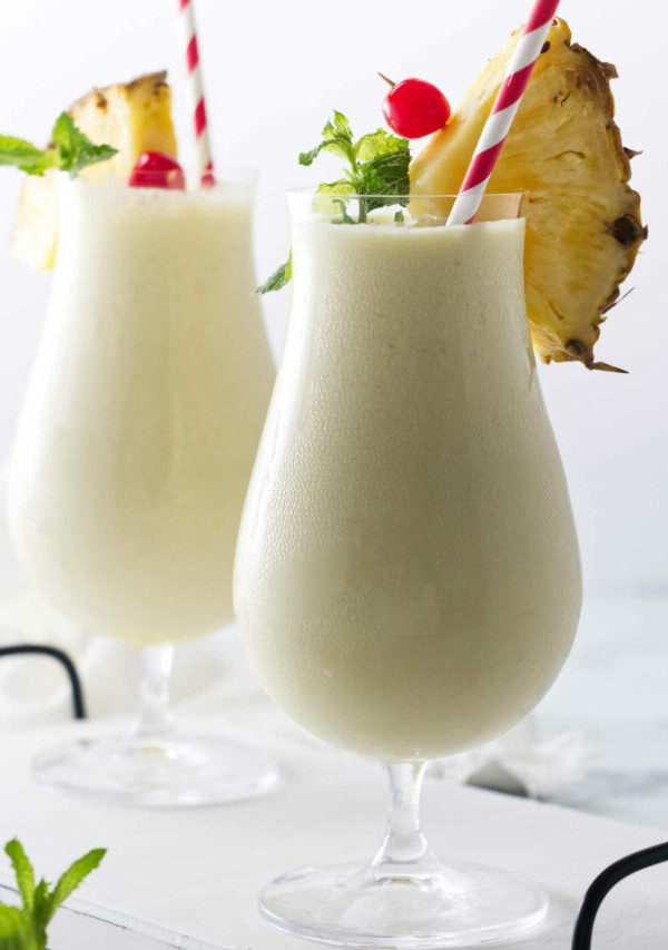 Two glasses of creamy non-alcohol beverage, with paper straws and garnishes of pineapple, cherry and mint.