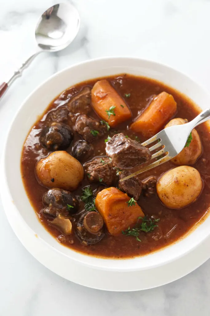 A serving of stew with meat, potatoes, mushrooms and carrots in a brown sauce. A dinner fork with a bite of meat.