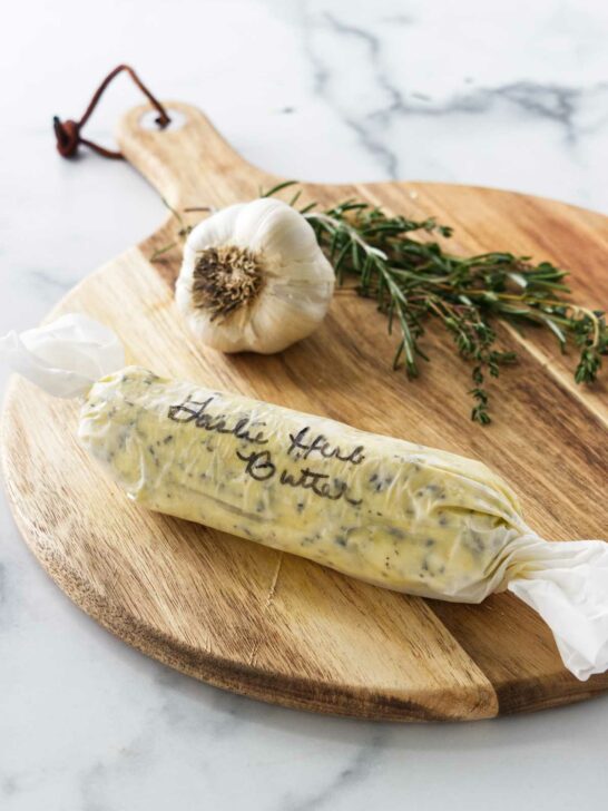 a cutting board with garlic, fresh hers and a wrapped log of compound butter.
