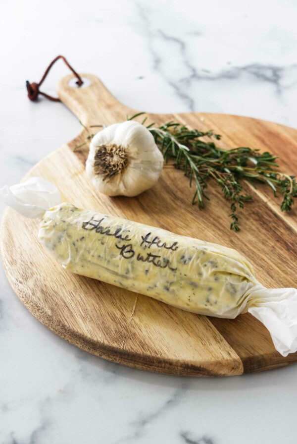 a cutting board with garlic, fresh hers and a wrapped log of compound butter.