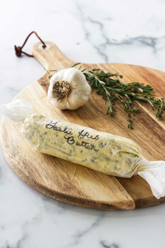 a cutting board with garlic, fresh hers and a wrapped log of compound butter.