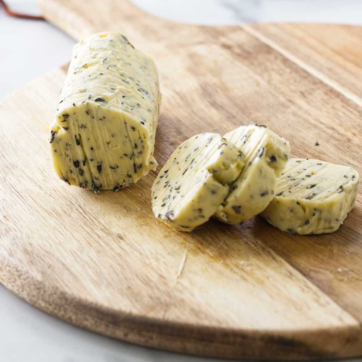 A roll of herbed butter on a cutting board with three slices from the butter.