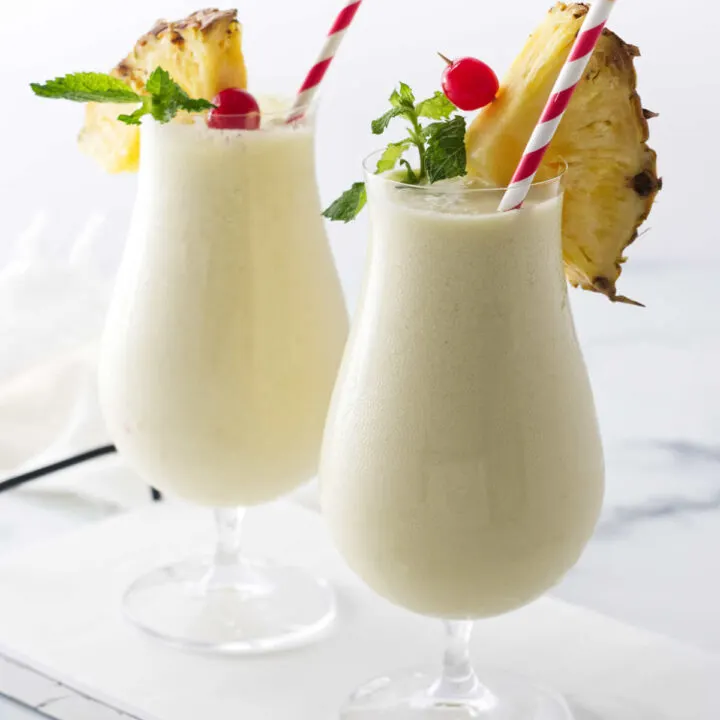 Two glasses of creamy non-alcohol beverage, with paper straws and garnishes of pineapple, cherry and mint.