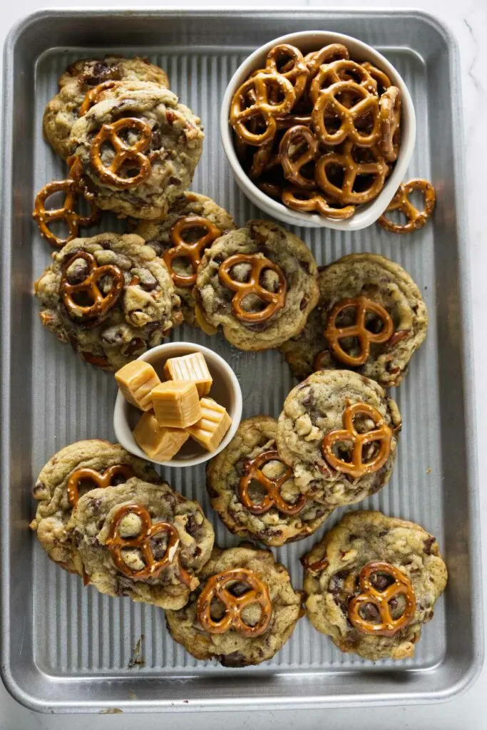 Several cookies loaded with pretzels and caramel next to dishes of caramel squares and miniature pretzels.