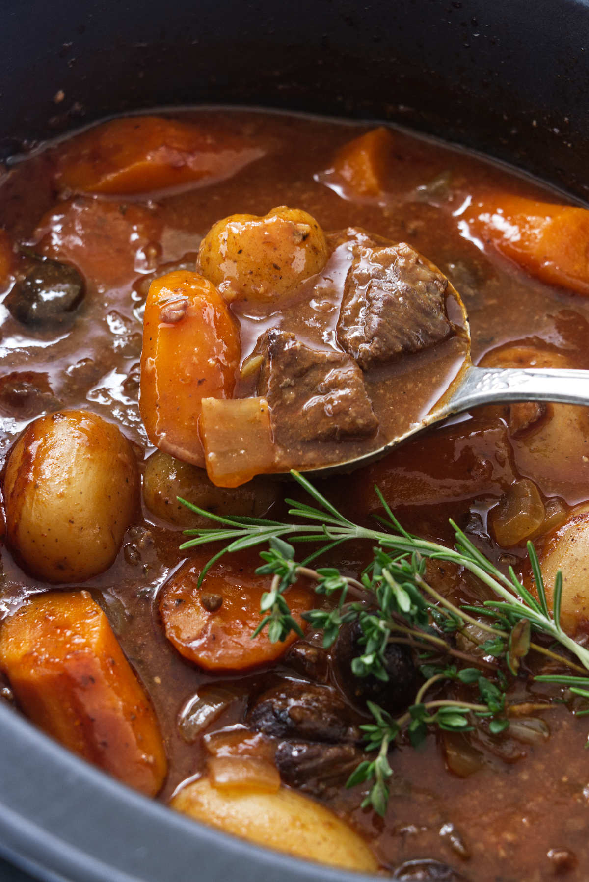 A close up photo of stew in a slow cooker. A large serving spoon filled with meat, potatoes and carrots.