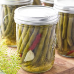 Pickled green beans in a canning jar with garlic and hot pepper, sitting on a cutting board with fresh dill in the corner.