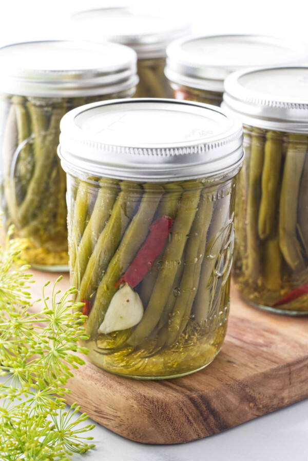 Pickled green beans in a canning jar with garlic and hot pepper, sitting on a cutting board with fresh dill in the corner.