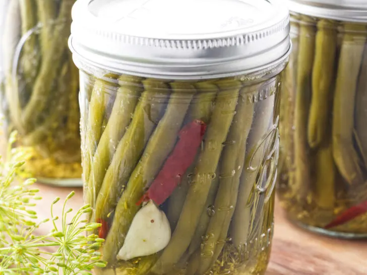 Pickled green beans in a canning jar with garlic and hot pepper, sitting on a cutting board with fresh dill in the corner.