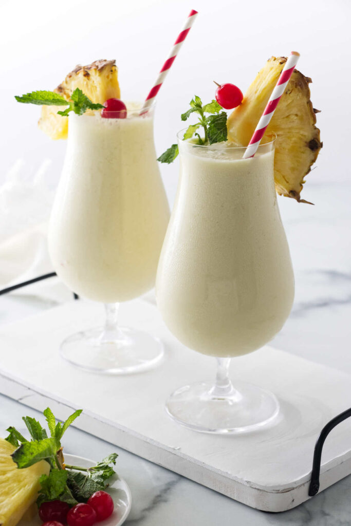Two tall hurricane glasses filled with beverage and garnished with pineapple wedges, cerries and mint.